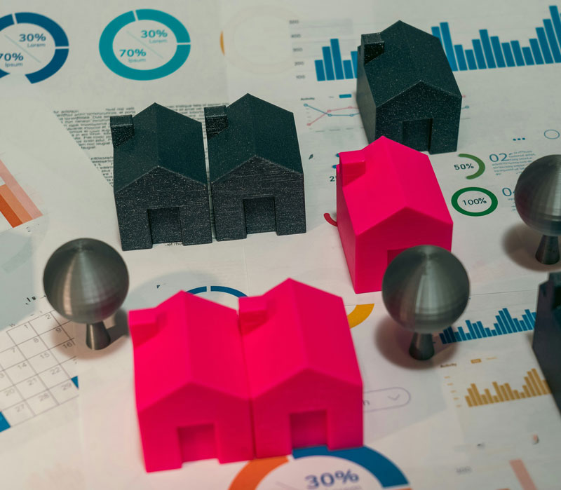 Small plastic game pieces in the shape of houses sit on top of paper reports showing a variety of types of charts and graphs.