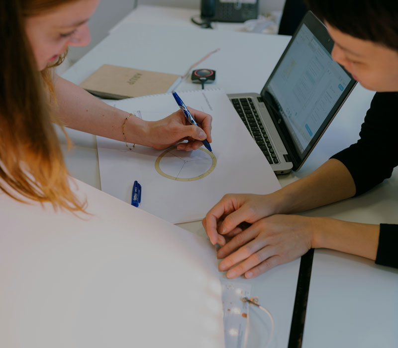 Two people drawing plans on a piece of paper. A laptop is open and on on the table behind them.